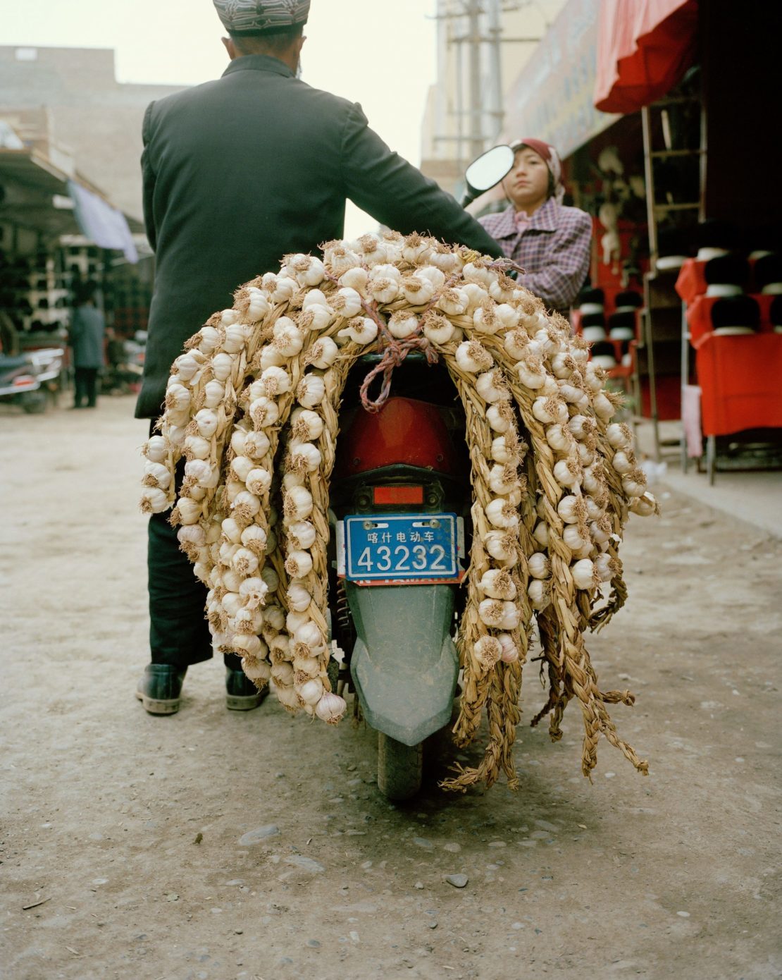 Man and woman riding motorcycle loaded with garlic in outdoor marketplace setting with hats for sale in the background.
