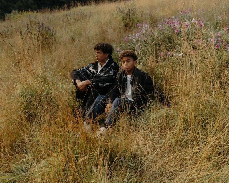 Two young men sit amongst tall grass in an outdoor setting, with a slight eye-level angle showcasing their casual attire and natural environment.