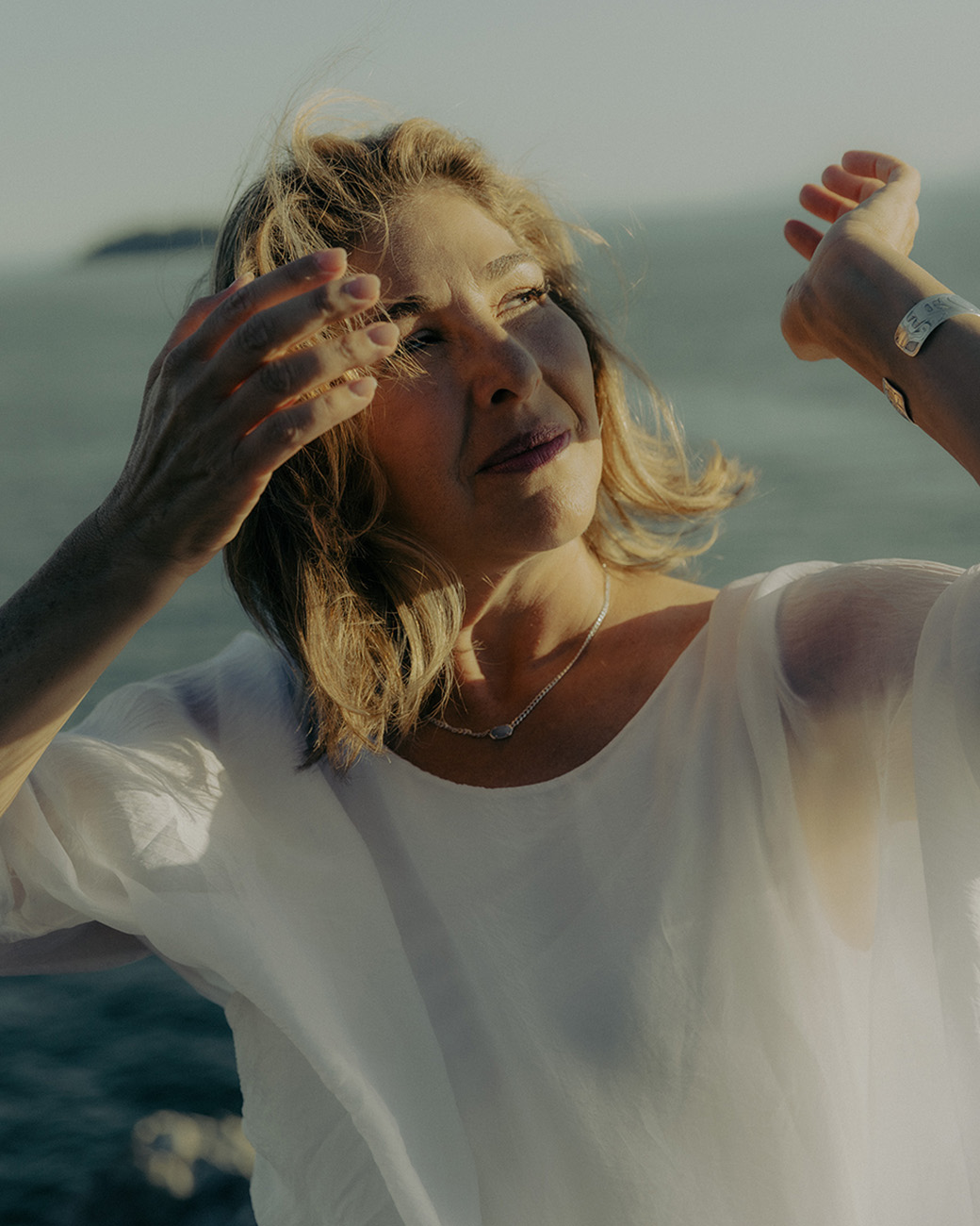 Mature woman in a white sheer top posing outdoors near the ocean with her hands raised and hair blowing.