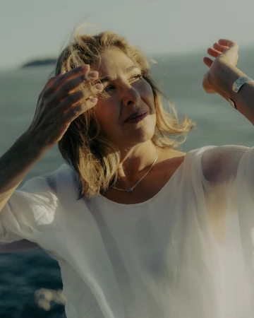 Mature woman in a white sheer top posing outdoors near the ocean with her hands raised and hair blowing.
