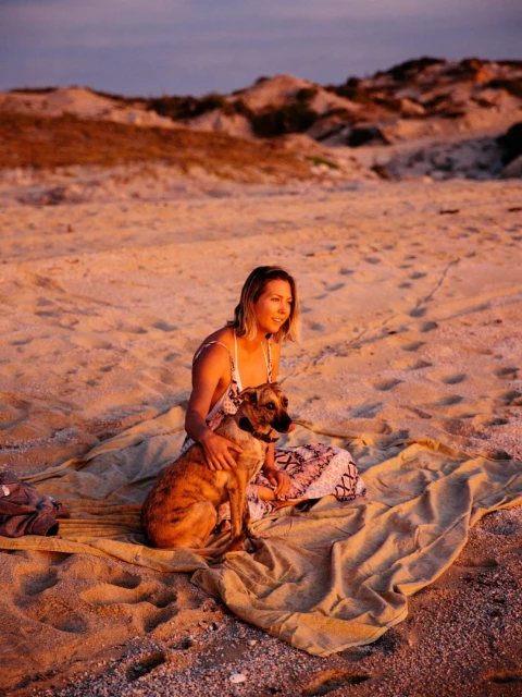 Woman sits with a dog on a blanket at the beach near dunes at sunset; the warm lighting and simple composition create a calm, natural scene.