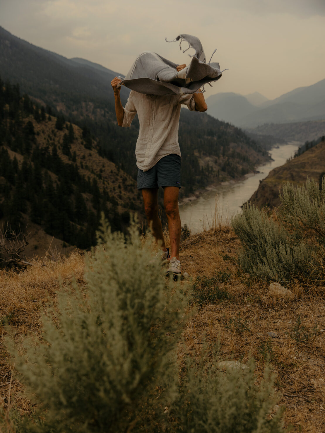 Person stands on a hillside near mountains and a river, holding a blanket. The shot captures nature and a sense of outdoor adventure.