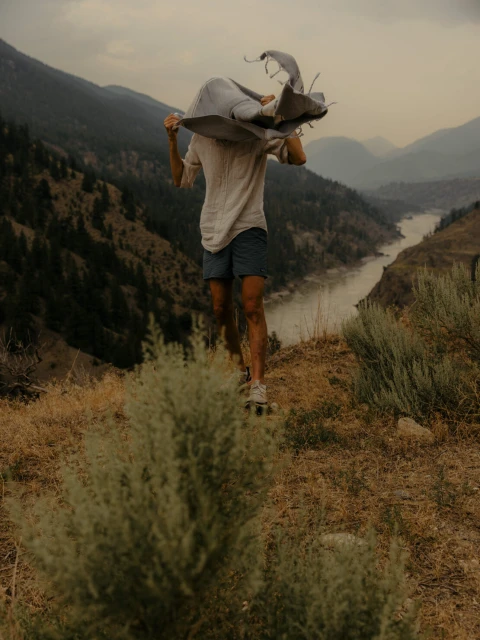 Person stands on a hillside near mountains and a river, holding a blanket. The shot captures nature and a sense of outdoor adventure.