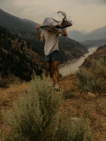Person stands on a hillside near mountains and a river, holding a blanket. The shot captures nature and a sense of outdoor adventure.