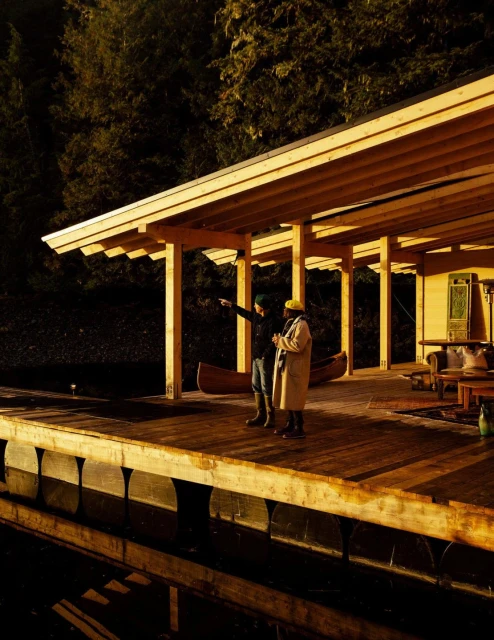 Two people stand under a covered wooden dock near a lake at night, with a canoe and wooden furniture visible in the background.