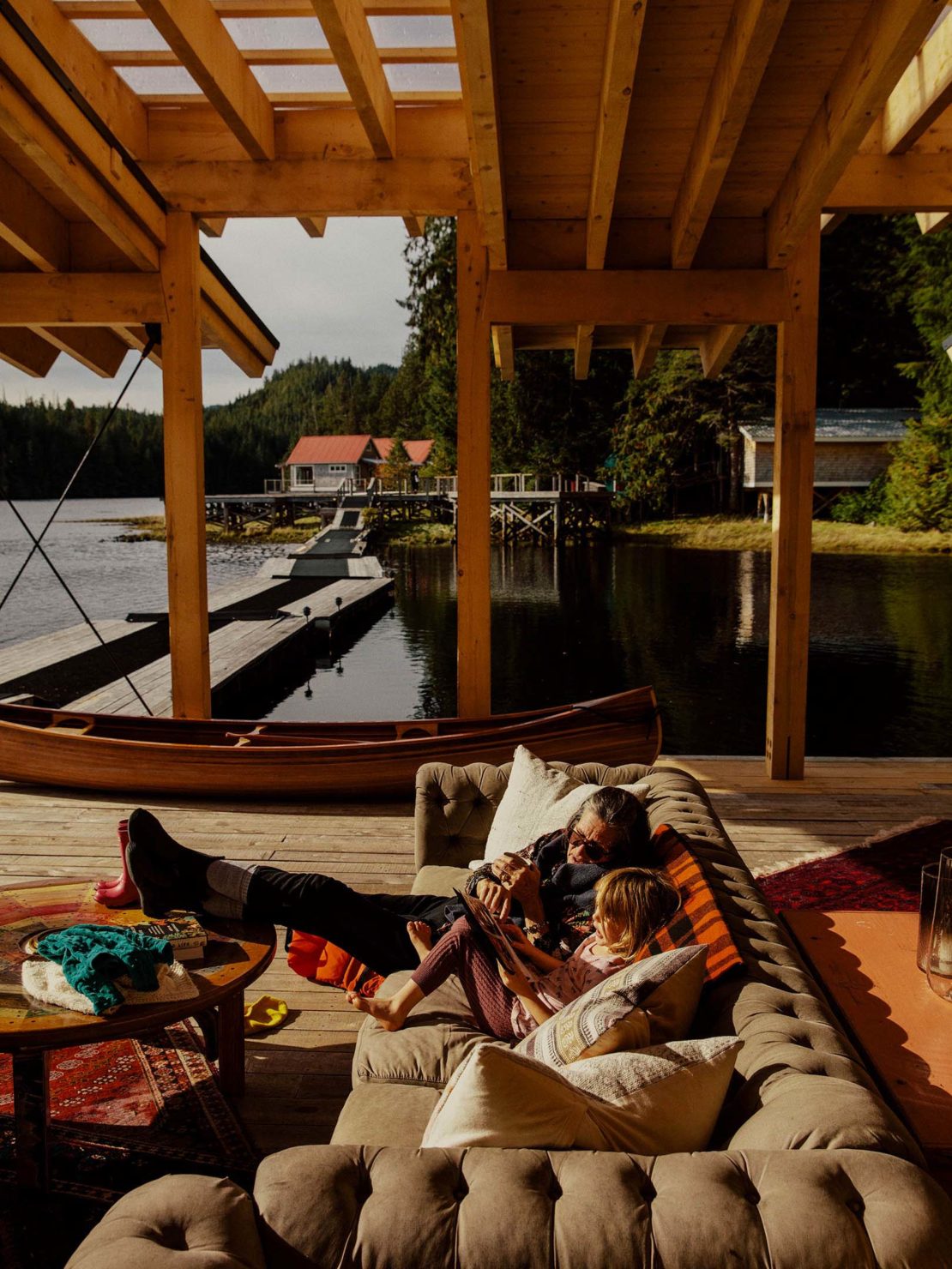 Father and daughter relaxing on an indoor/outdoor sofa on a wooden deck overlooking a lake with forest and buildings on docks.