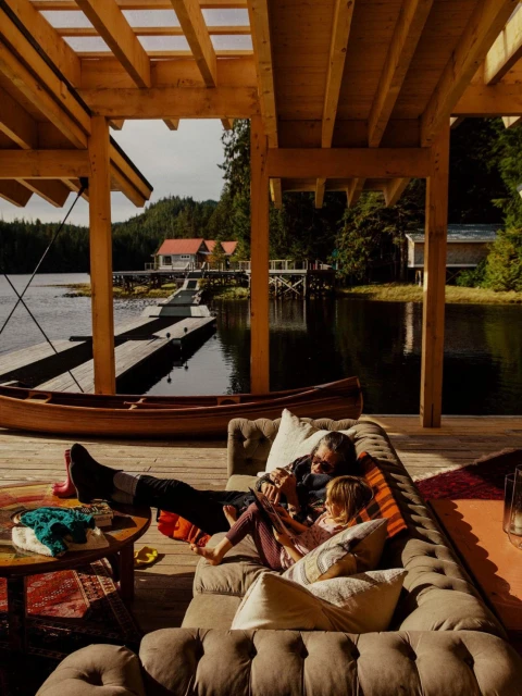 Father and daughter relaxing on an indoor/outdoor sofa on a wooden deck overlooking a lake with forest and buildings on docks.