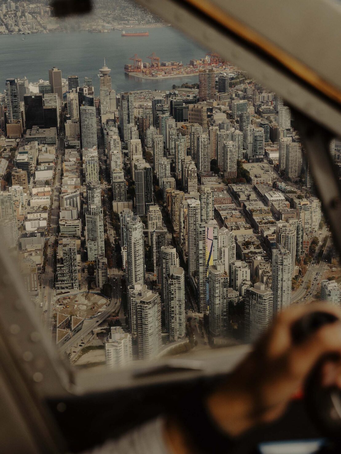Aerial cityscape view from airplane window, showing high-rise buildings, waterfront with cargo port, mountains in background on overcast day.