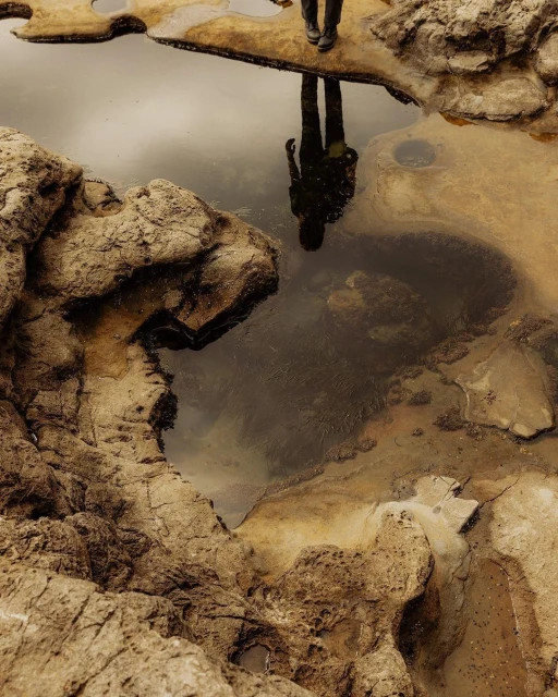 Adult stands on rocky outcrop by a pool of water, their reflection visible in the water, surrounded by textured rocks.