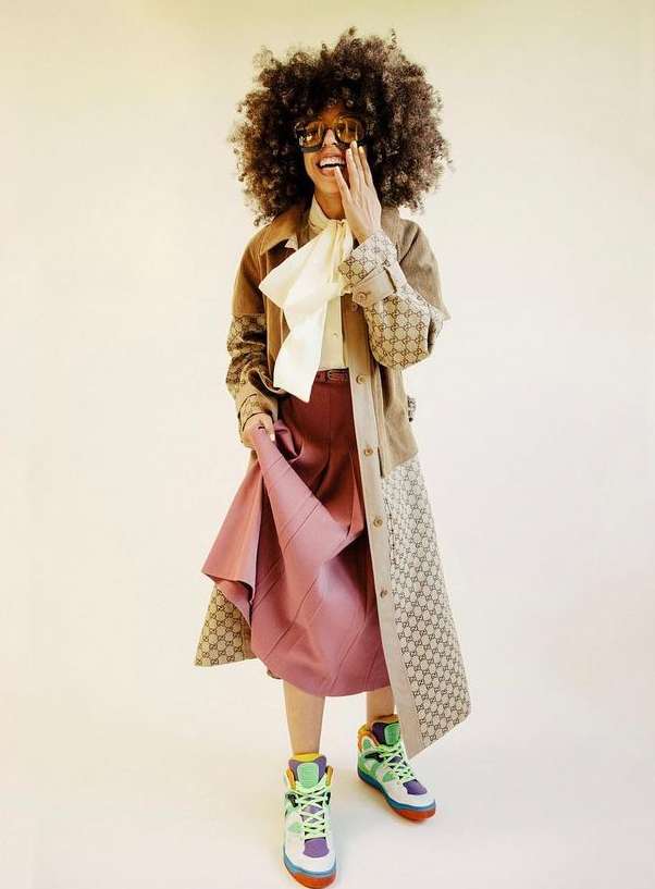 Stylish African American woman poses in vintage-inspired Gucci outfit, featuring a tan jacket, silk blouse, and a line skirt on beige.