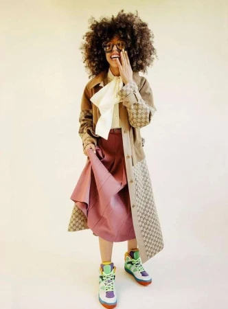 Stylish African American woman poses in vintage-inspired Gucci outfit, featuring a tan jacket, silk blouse, and a line skirt on beige.
