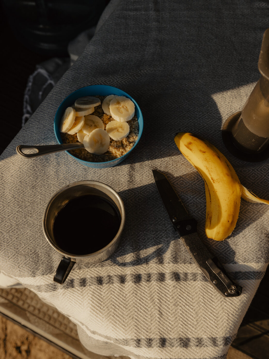 Breakfast scene: a blue bowl of sliced bananas and oatmeal, a metal mug of coffee, a knife, and a whole banana on a patterned cloth.
