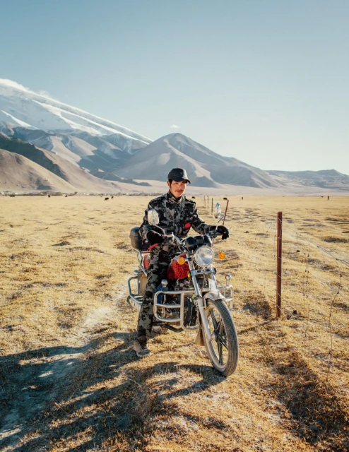 Adult male in camouflage rides a motorcycle in a sunny grassland plain with snowy mountains in the background against a blue sky.