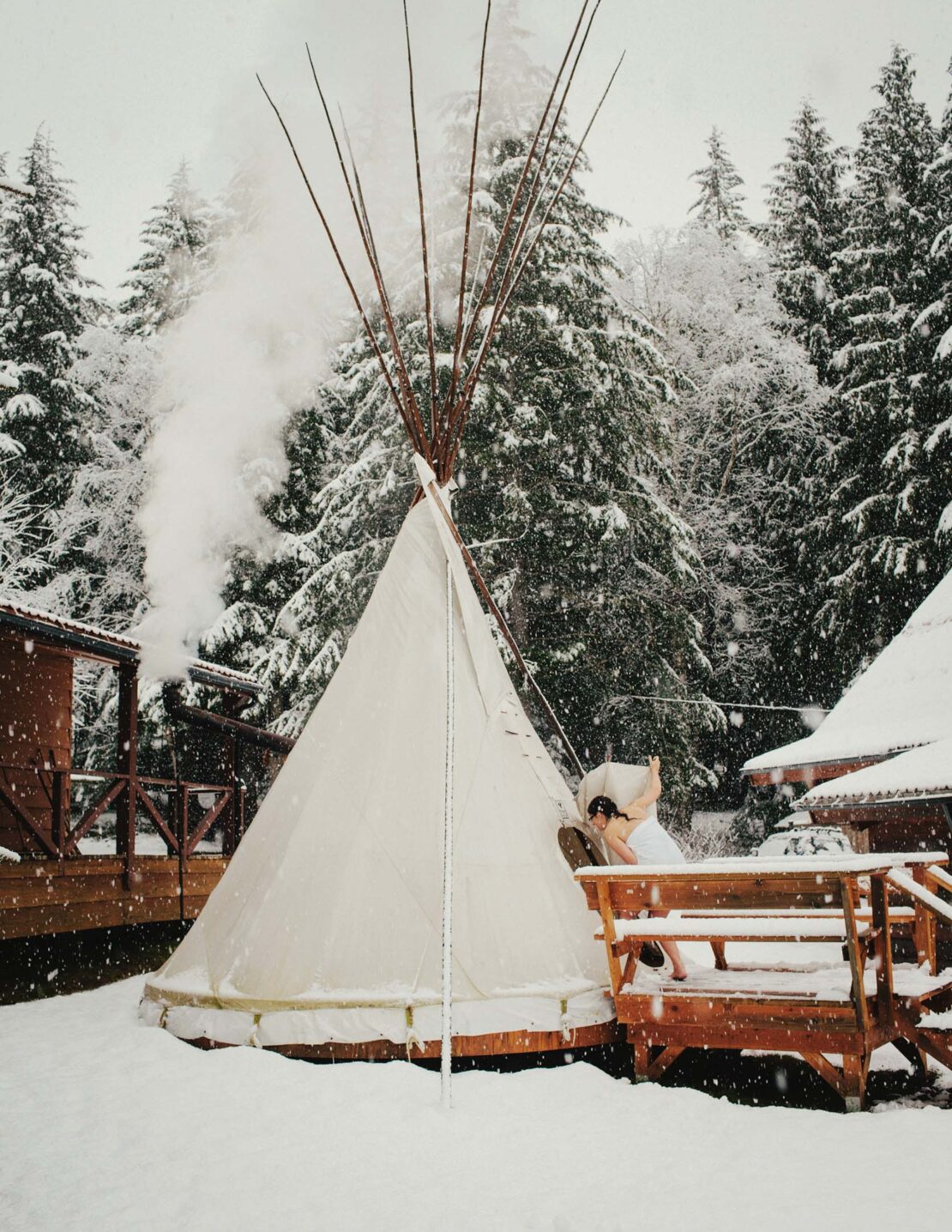 A tipi stands in a snowy landscape with trees and buildings in the background, creating a serene winter scene.  