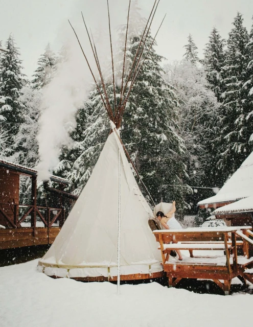 A tipi stands in a snowy landscape with trees and buildings in the background, creating a serene winter scene.  