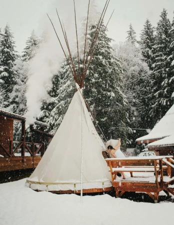 A tipi stands in a snowy landscape with trees and buildings in the background, creating a serene winter scene.  