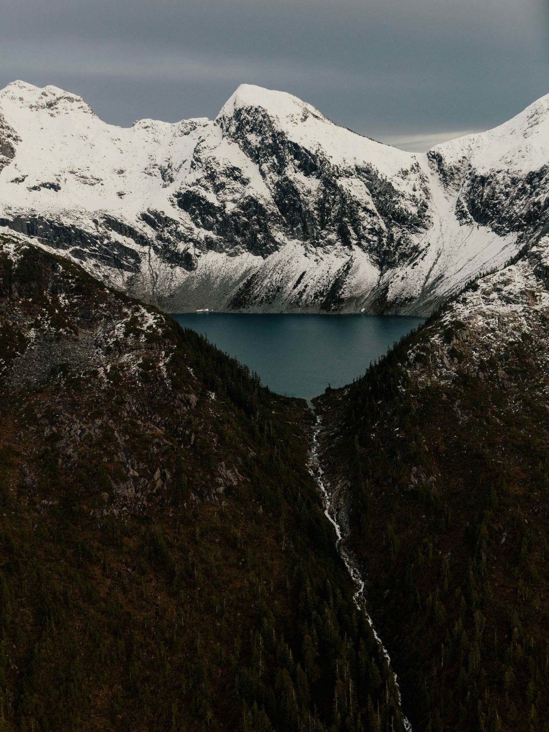 Scenic view of a lake surrounded by mountains covered in snow, with a small stream flowing down a forested slope in the foreground.