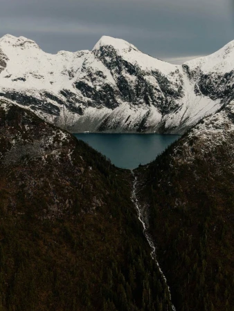Scenic view of a lake surrounded by mountains covered in snow, with a small stream flowing down a forested slope in the foreground.