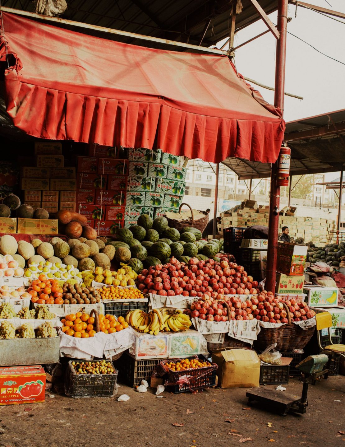 Produce stall at an outdoor market with a variety of fruits and vegetables displayed under a red awning on tiered levels and boxes.
