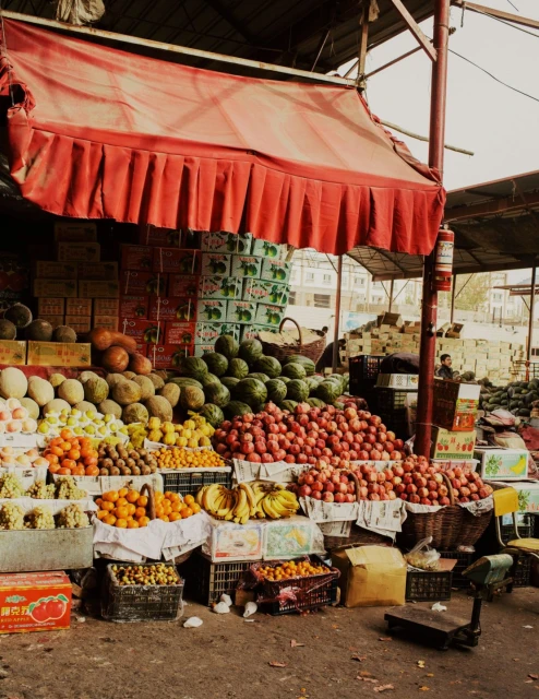 Produce stall at an outdoor market with a variety of fruits and vegetables displayed under a red awning on tiered levels and boxes.