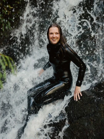 Smiling woman wearing a black wetsuit seated on rocks beneath a rushing waterfall photographed by Grant Harder