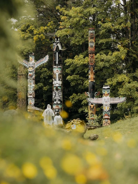 Several totem poles with colorful carvings stand on a grassy hill in a park-like setting, with two people in raincoats in the background.