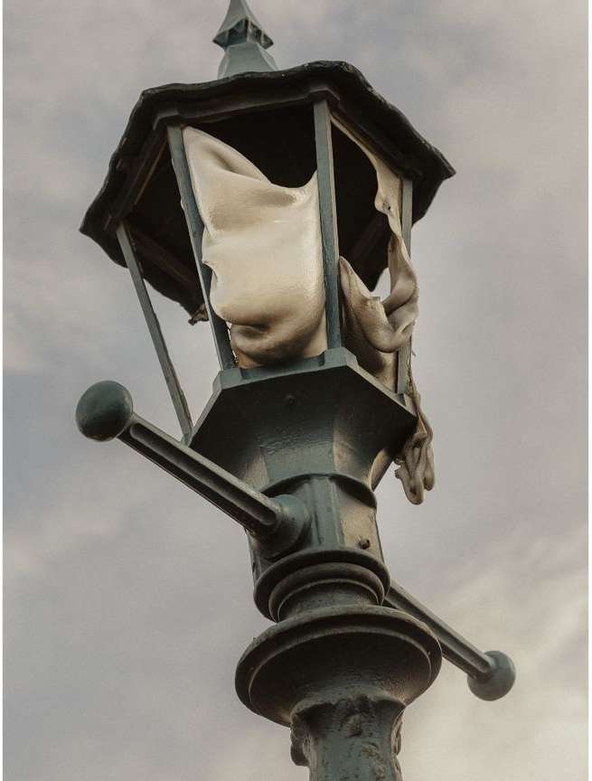 Artistic image of an antique-style street lamp with torn fabric inside, set against a cloudy sky, evoking a sense of decay and mystery.