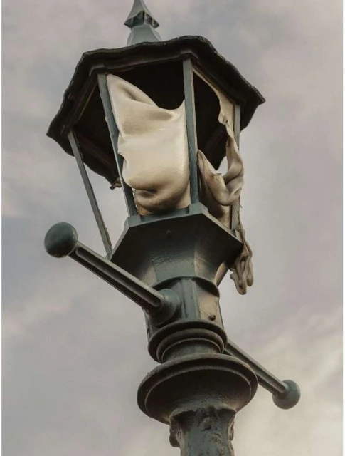 Artistic image of an antique-style street lamp with torn fabric inside, set against a cloudy sky, evoking a sense of decay and mystery.