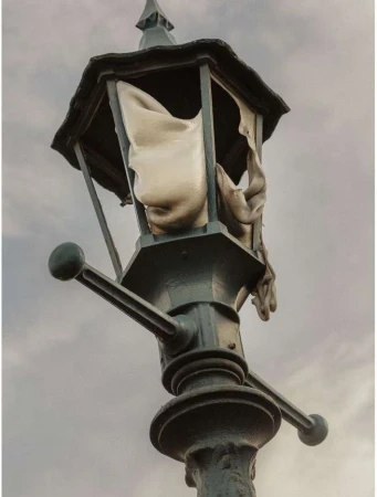 Artistic image of an antique-style street lamp with torn fabric inside, set against a cloudy sky, evoking a sense of decay and mystery.