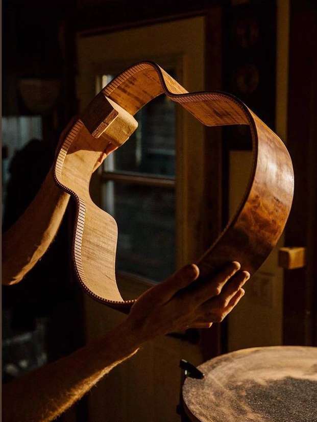 Craftsperson holds curved, unfinished wooden guitar body in a dimly lit workshop with woodworking tools. Backlit by a nearby window.