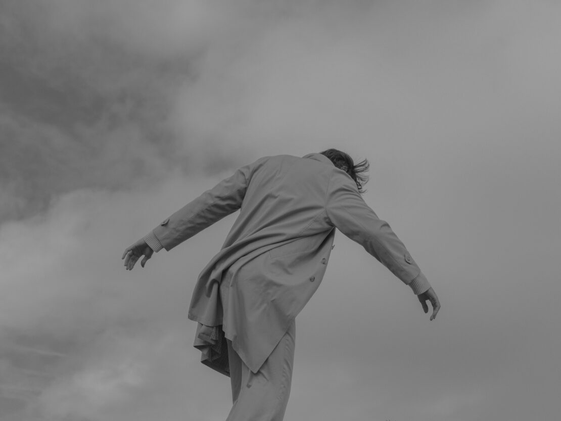 Monochromatic low angle shot of adult with blowing hair, wearing a light-colored coat against a cloudy sky.