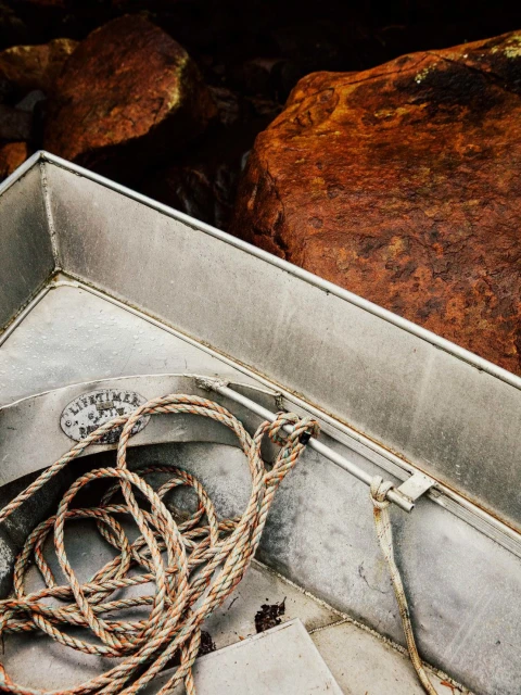 Close-up of a weathered metal dinghy filled with a coiled brown rope, set against a backdrop of dark, moss-covered boulders.