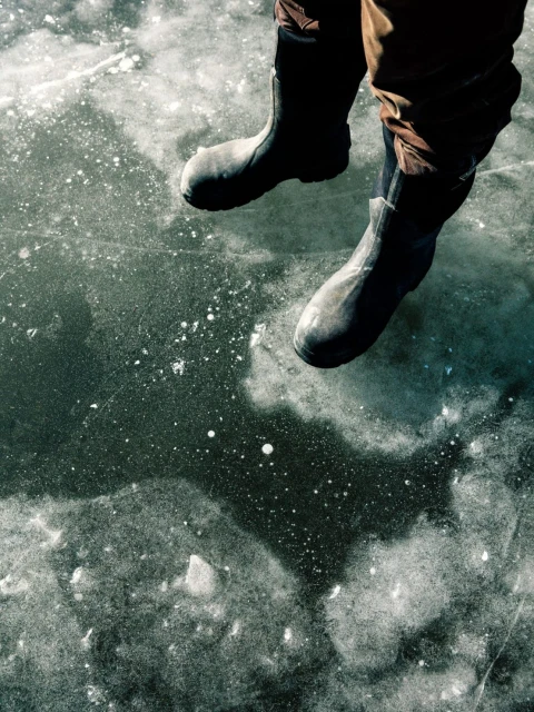 Overhead shot of a person wearing black boots sitting on a frozen lake with cracked ice and air bubbles visible.