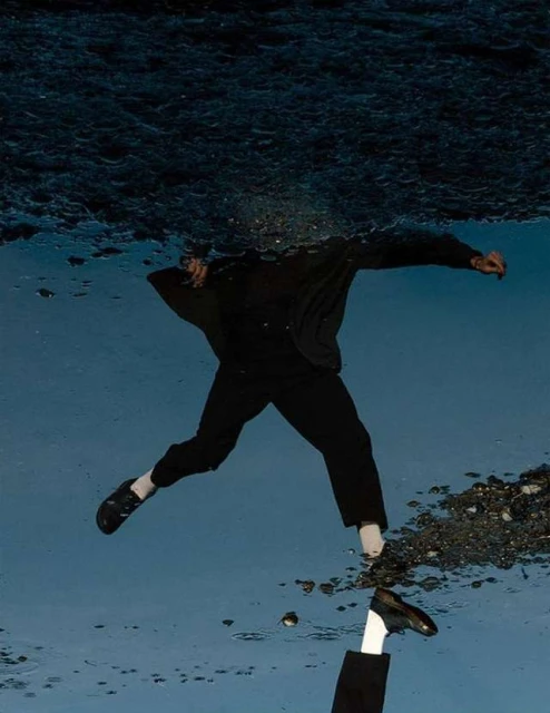 Reflection of a person in black clothing walking on a wet surface, shoes and socks are visible in the water reflection.