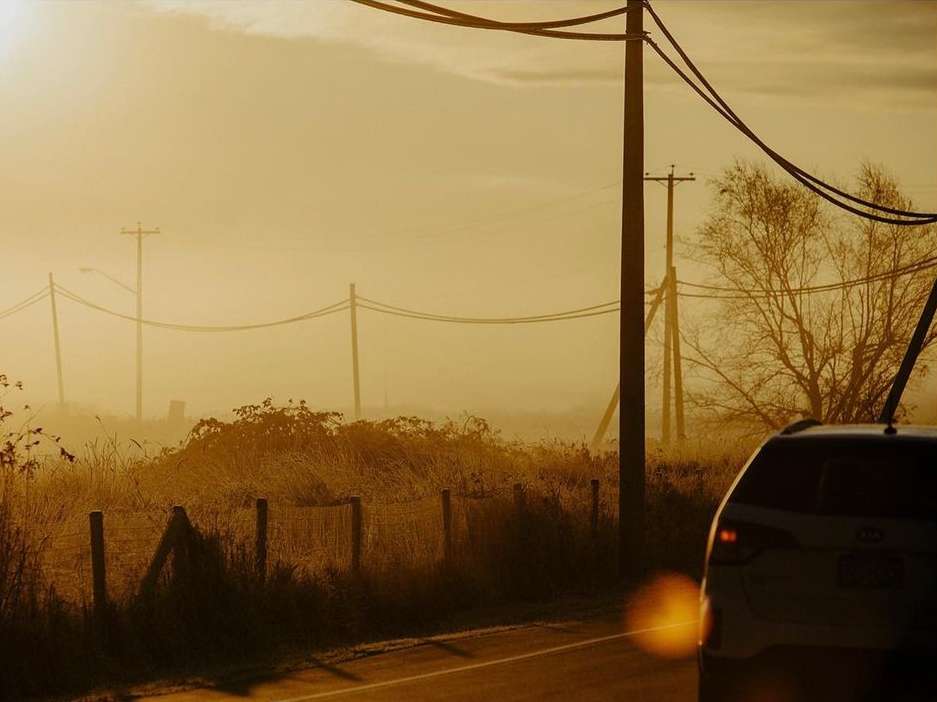 Car on asphalt road with utility poles, vegetation, and heavy fog creating golden-colored scenic landscape at sunrise or sunset.