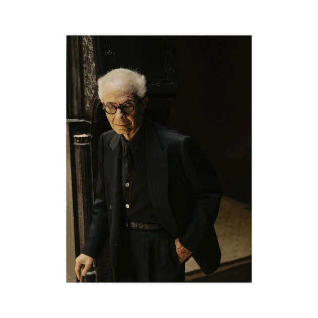 Older adult male in a dark suit and glasses, with short white hair, stands indoors with a cane, set against a dark background.