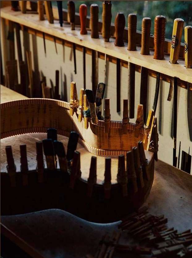 Close-up of a partially assembled wood guitar in a workshop with wooden pegs and various tools arranged in the background.