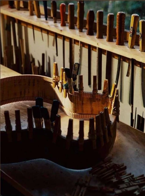 Close-up of a partially assembled wood guitar in a workshop with wooden pegs and various tools arranged in the background.