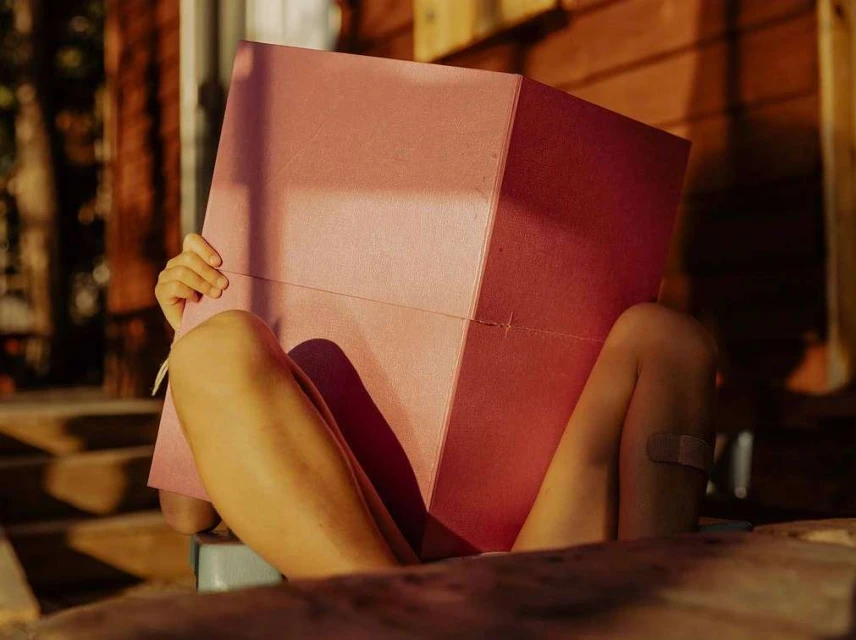 A child sits holding a pink box in front of their face, partially obscuring it, outdoors with the warm tones of wooden architecture.