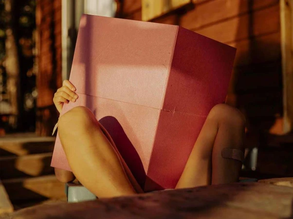 A child sits holding a pink box in front of their face, partially obscuring it, outdoors with the warm tones of wooden architecture.
