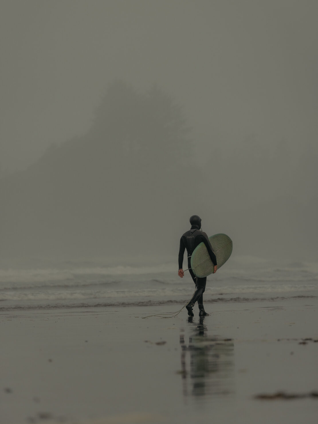 Surfer in a black wetsuit walking along a wet sandy beach while carrying a surfboard under a heavy fog.