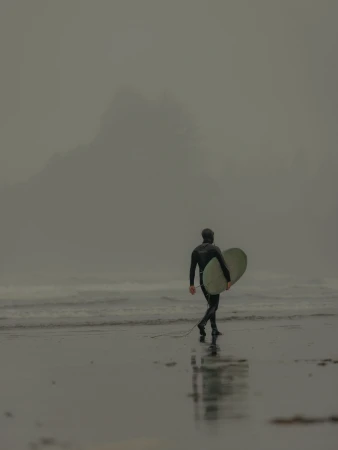 Surfer in a black wetsuit walking along a wet sandy beach while carrying a surfboard under a heavy fog.