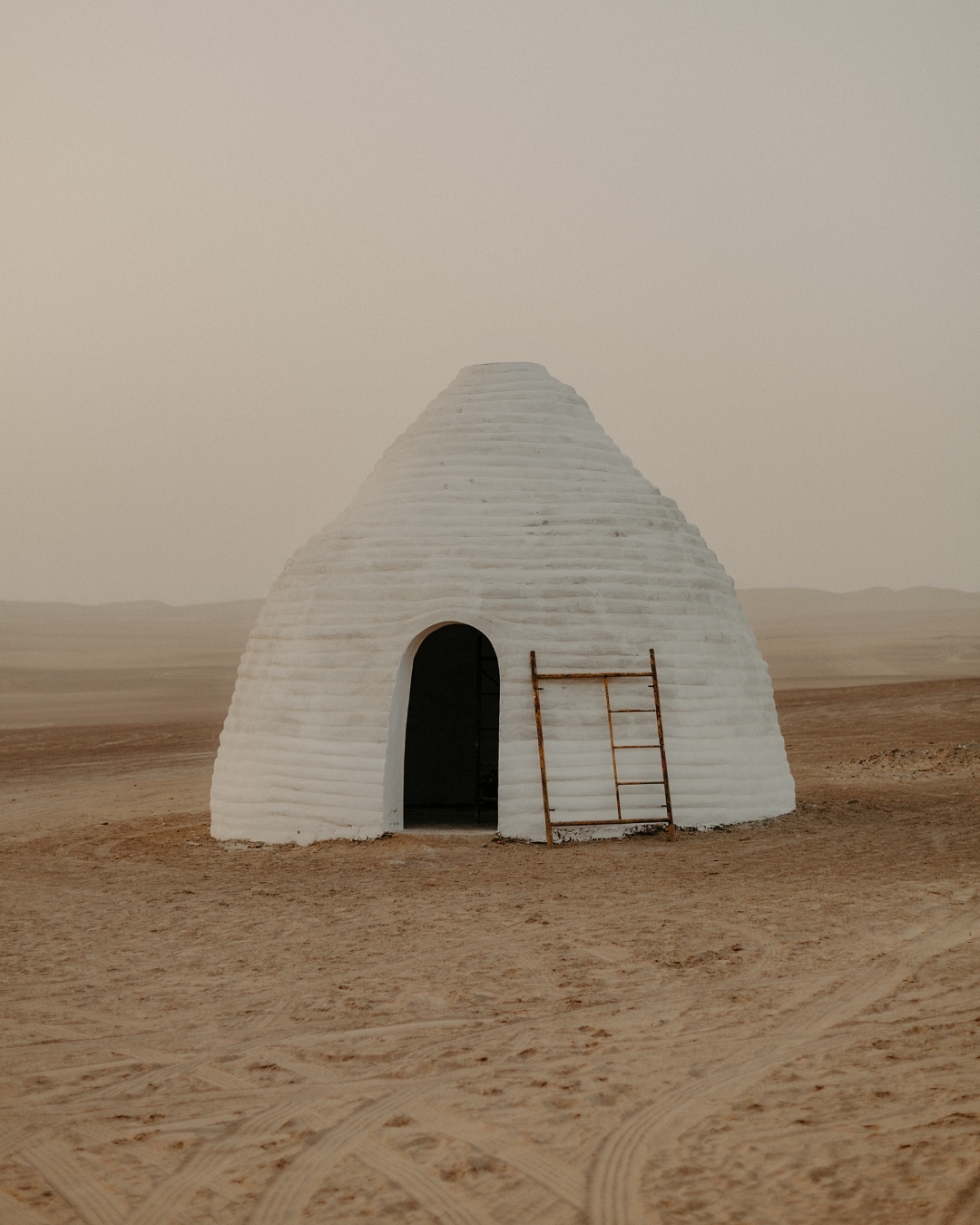 White domed building with a ladder in the desert under a hazy sky. The sandy landscape shows tire tracks near the structure.