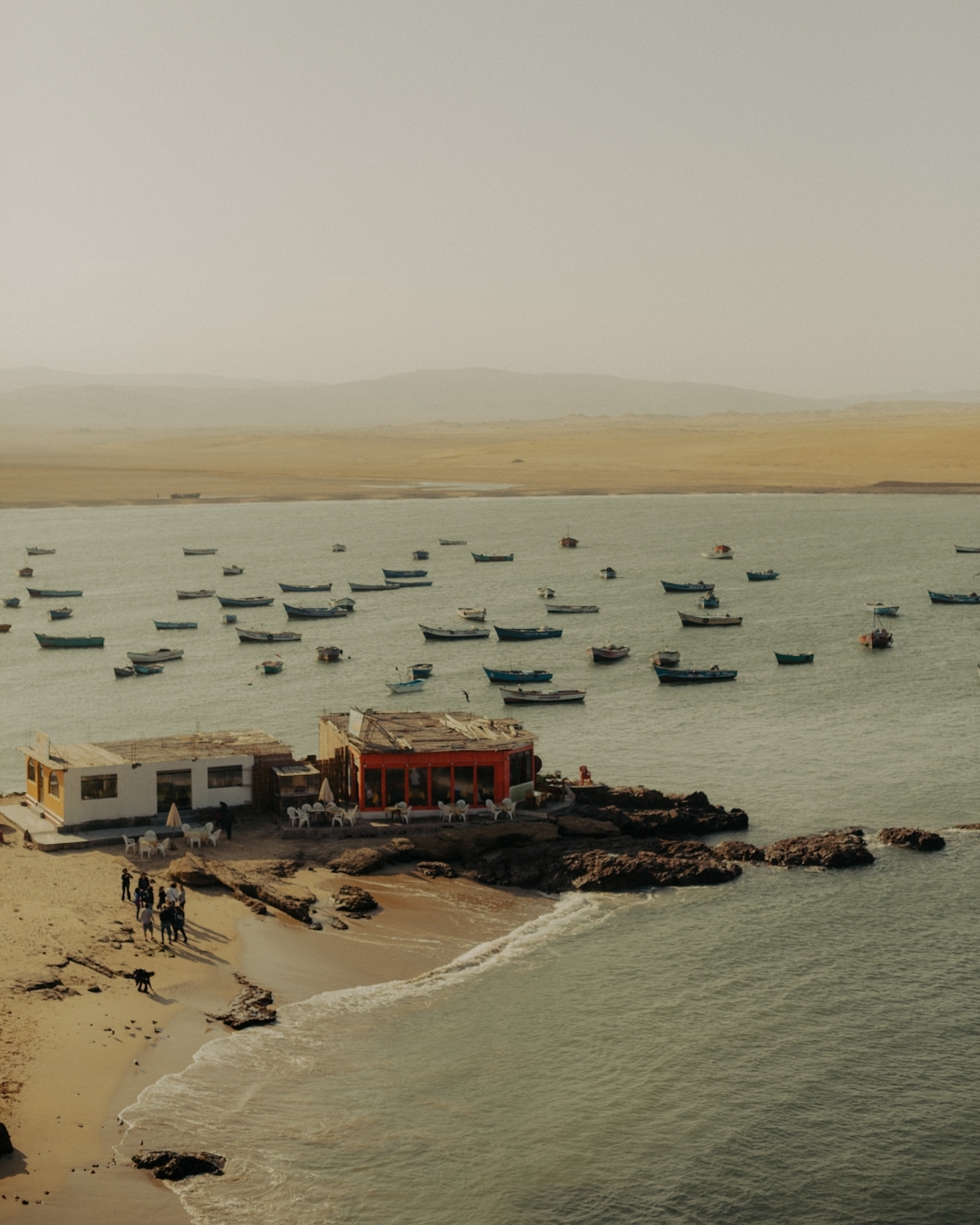 Scenic view of boats on the water near a sandy beach, with buildings, people, and a mountainous landscape in the background.