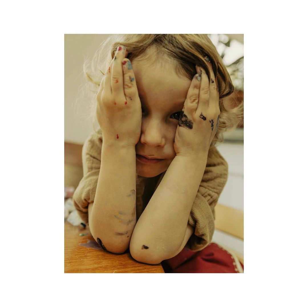 Close-up of a child with hands on their face, hands and forearms covered in dark paint, sitting at a wooden table in an indoor setting.