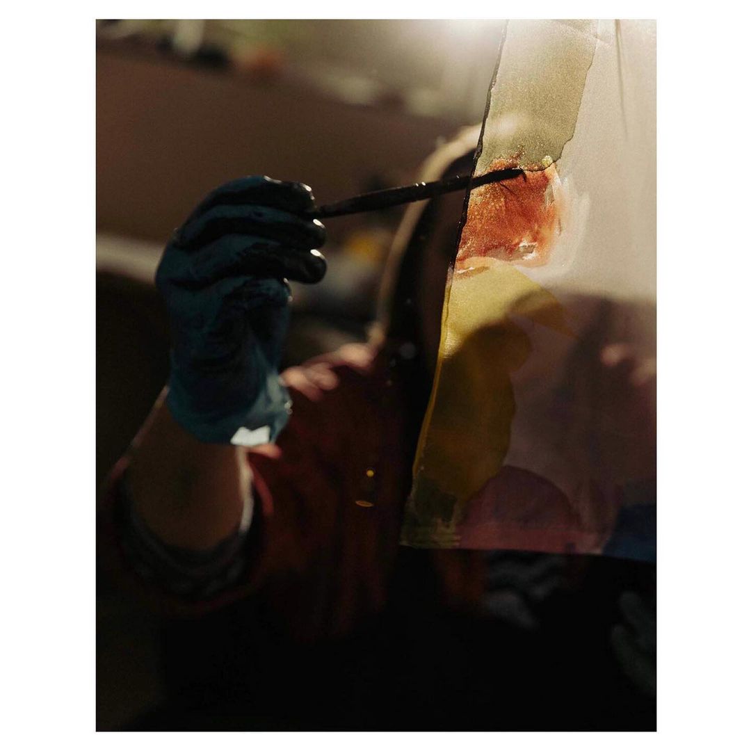 Woman painting on transparent material indoors. She wears blue gloves and a red shirt while applying color to a clear surface.