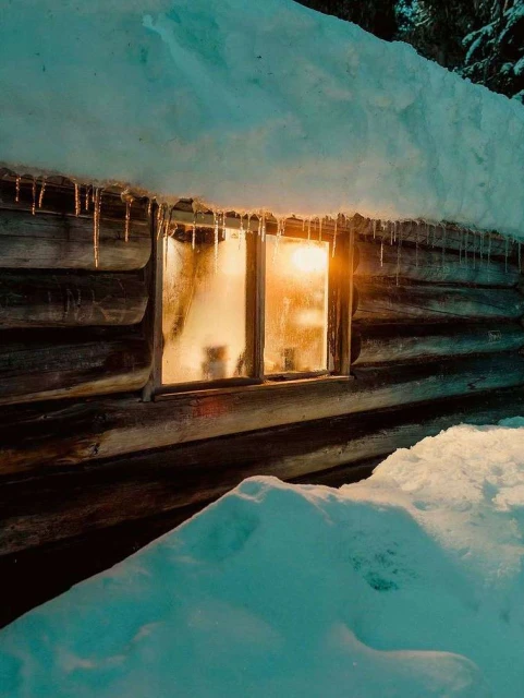 Rustic wooden cabin covered in snow during winter with a window reflecting warm light, evoking a cozy and secluded atmosphere.