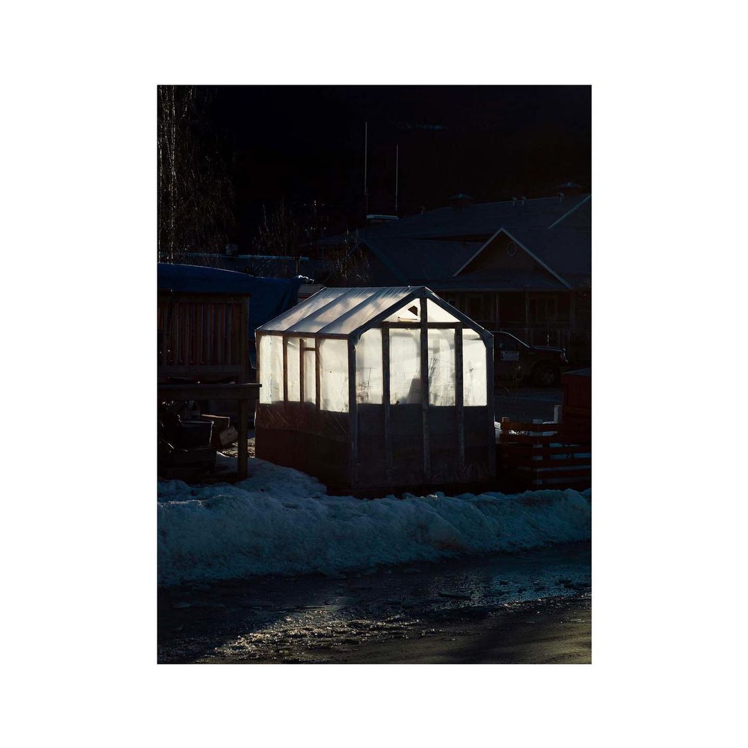 Illuminated greenhouse on a snowy ground in a rural setting at dusk with houses visible in the dark background, creating a peaceful atmosphere.