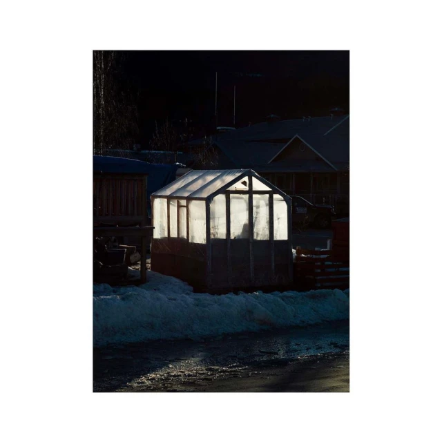Illuminated greenhouse on a snowy ground in a rural setting at dusk with houses visible in the dark background, creating a peaceful atmosphere.