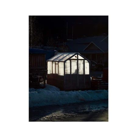 Illuminated greenhouse on a snowy ground in a rural setting at dusk with houses visible in the dark background, creating a peaceful atmosphere.